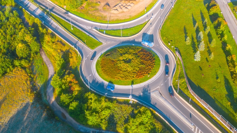 Aerial perspective of a busy roundabout with multiple exits, surrounded by lush green trees and landscape, bathed in sunlight.