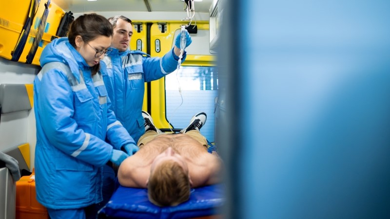 One of two young paramedics examining unconscious shirtless man on stretcher while her colleague holding dropper