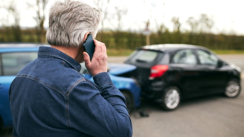 Rear view of senior male driver standing by damaged car after traffic accident reporting incident