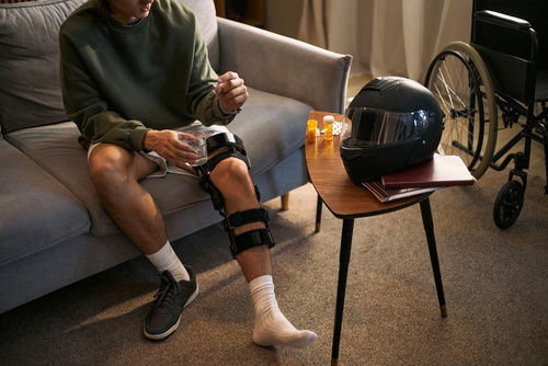 injured man wearing knee brace seated on couch with motorcycle helmet and wheelchair nearby